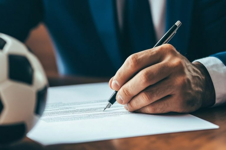 A man signing a piece of paper, with a mini football on  the table in front of him.